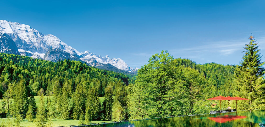 Infinitypool, in dem sich grüner Wald und Bergpanorama spiegeln, am Rande drei Sonnenliegen