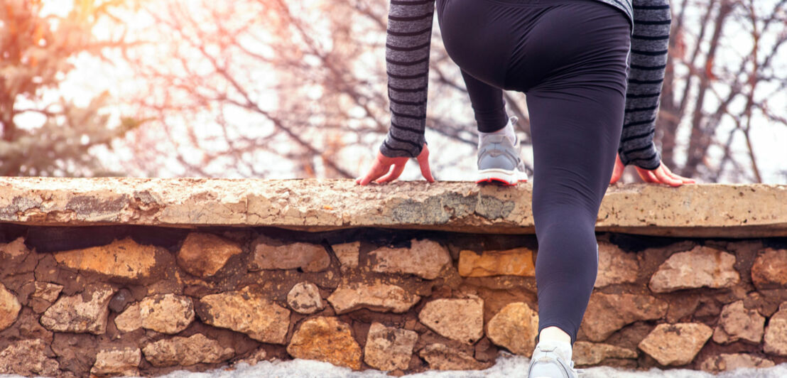 Jogger:in von hinten beim Dehnen der Wade an einem Mäuerchen