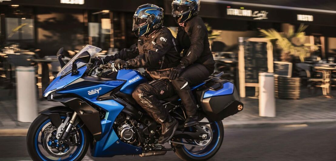 Eine blaue Suzuki GSX-S1000GT mit Fahrer und Sozia rollt vor einem Bistro vorbei