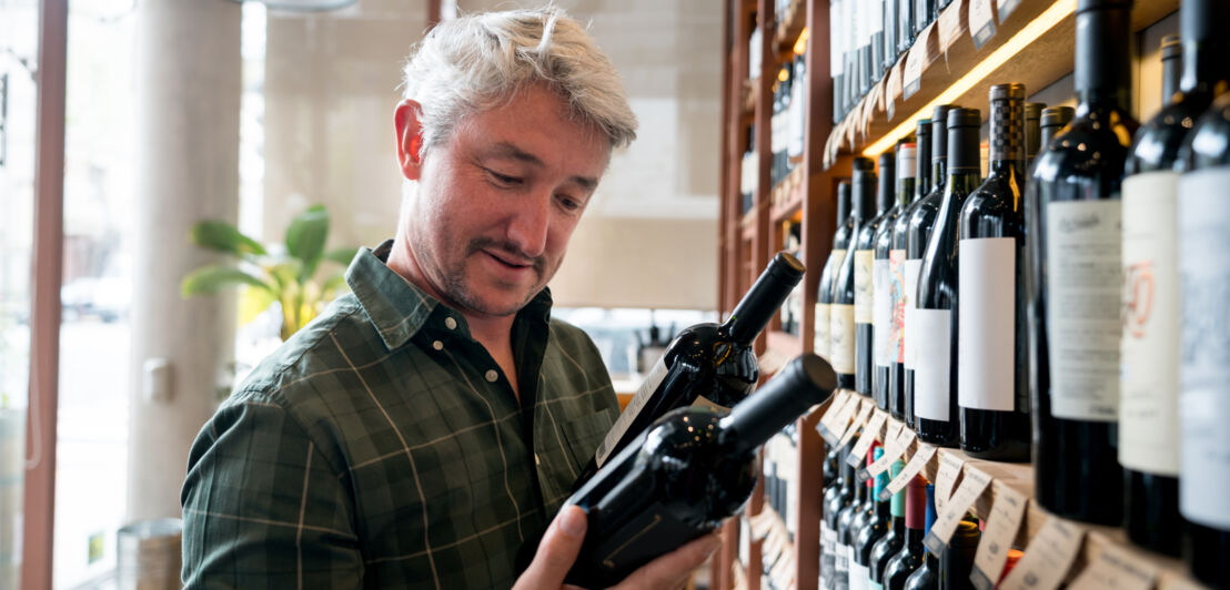 Ein Mann begutachtet zwei Weinflaschen vor einem Regal voller Wein.