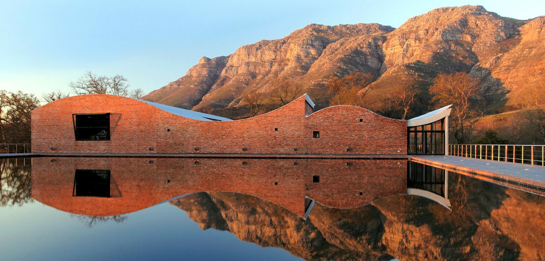 Modernes Gebäude aus rotem Stein mit geschwungenem Dach, das sich in einem Wasserbecken spiegelt, im Hintergrund ein Gebirge in der Abendsonne