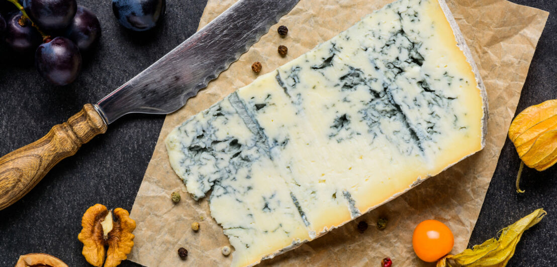 Ein Stück Blauschimmelkäse auf einem braunen Papier, garniert mit Nüssen und Früchten sowie einem Käsemesser