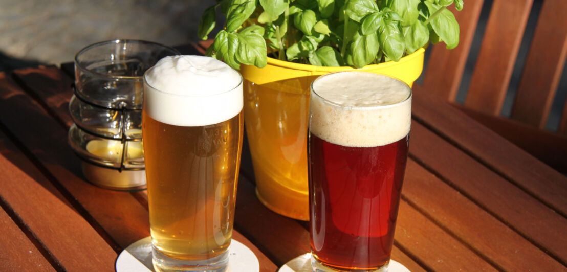 Ein dunkles und ein helles Bier in Gläsern stehen auf einem Gartentisch aus Holz