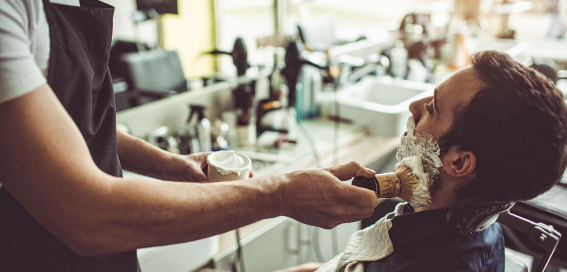 Ein Mann sitzt in einem Friseursessel und wird von seinem Barbier mit Rasierschaum eingepinselt