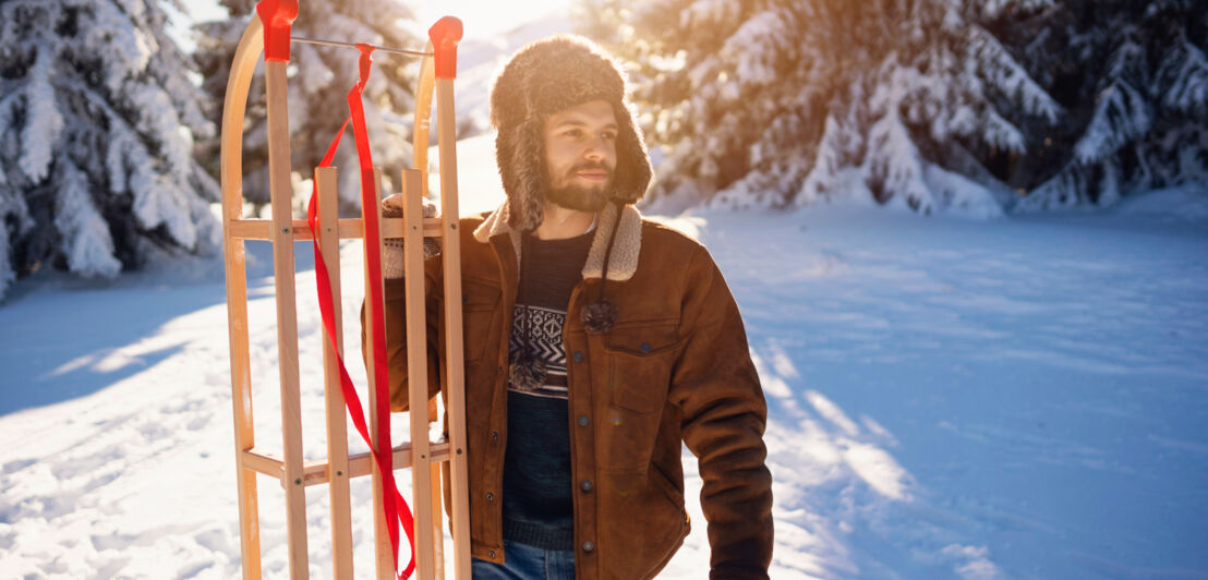 Ein Mann in Winterkleidung mit einem Schlitten in schneebedeckter Waldlandschaft.