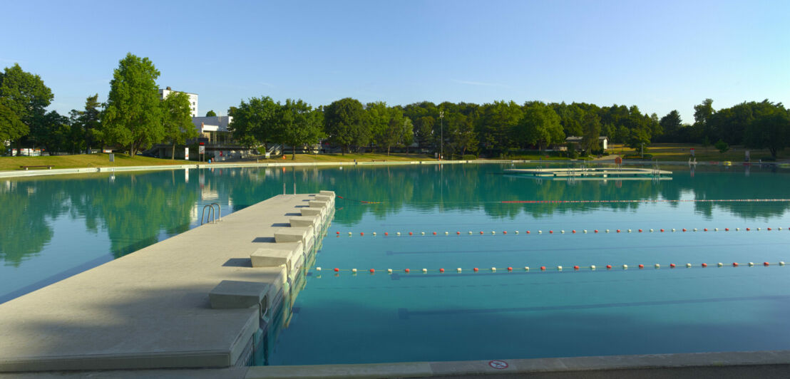 Ein Baggersee-ähnliches Schwimmbecken im Freien, mit abgetrennten Bahnen und einer schmalen, ringförmigen Kunstinsel.