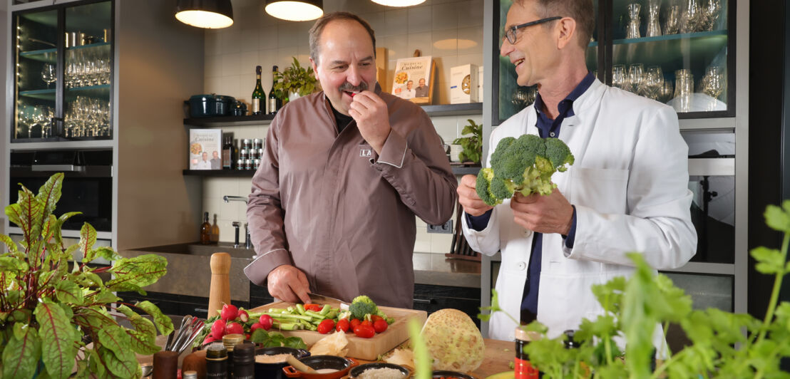 Starkoch Johann Lafer und Ernährungsmediziner Dr. Matthias Riedl stehen nebeneinander in einer Küche vor einer Theke mit rohem Gemüse