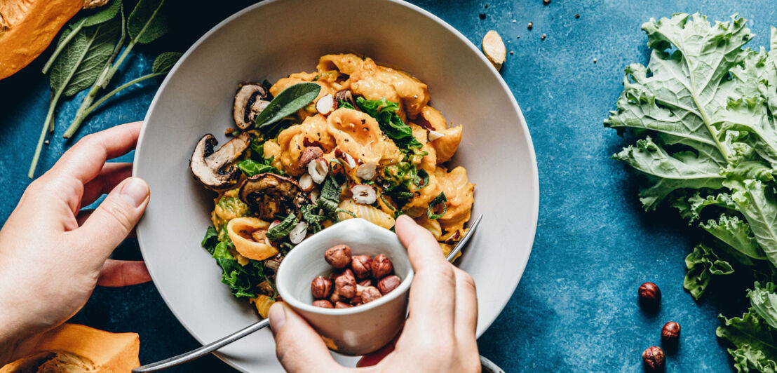 Eine Hand schüttet Haselnüsse auf ein Nudelgericht mit Kürbis und Champignons