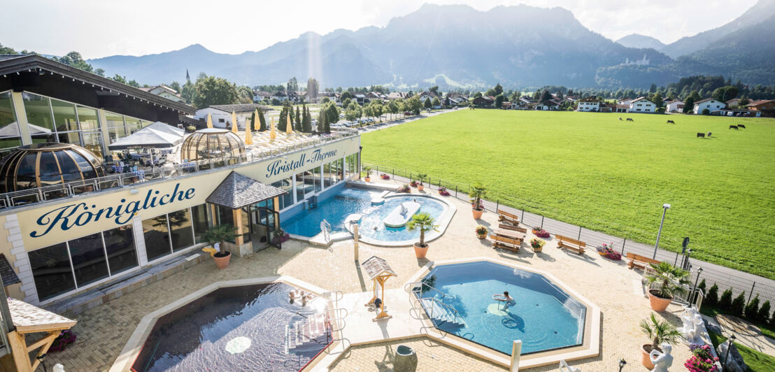 Eine Badelandschaft mit Terrasse und drei Wasserbecken, im Hintergrund grüne Berglandschaft mit Schloss Neuschwanstein