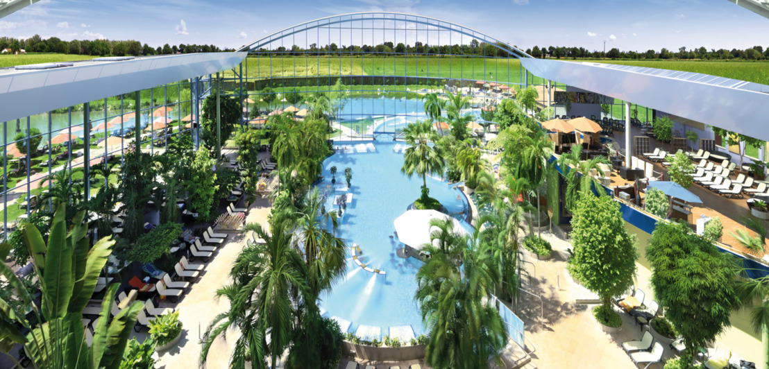 Panoramasicht auf eine große Badelandschaft mit einem großem Schwimmbecken und tropischen Pflanzen unter einer geöffneten Kuppel, die den Blick auf den blauen Himmel freigibt