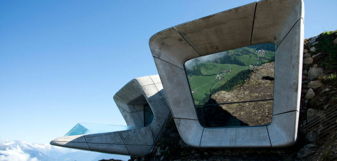 Zwei Betonauswüchse eines Gebäudes ragen aus einem Berg, im Hintergrund Berge