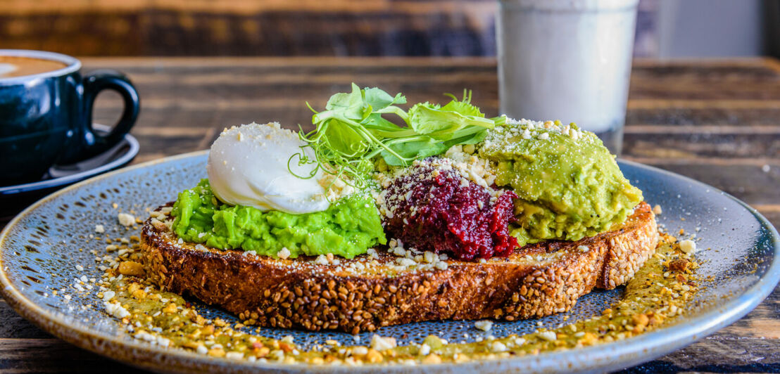 Ein Brot mit Guacamole, Ei und Rohkost auf einem blauen Teller auf einem Holztisch