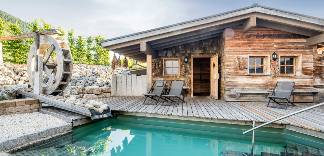 Ein kleines Holzhaus mit Pool davor, rechts daneben eine alte Mühle