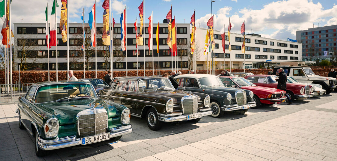 Oldtimer in einer Reihe vor einem Gebäude und Fahnen