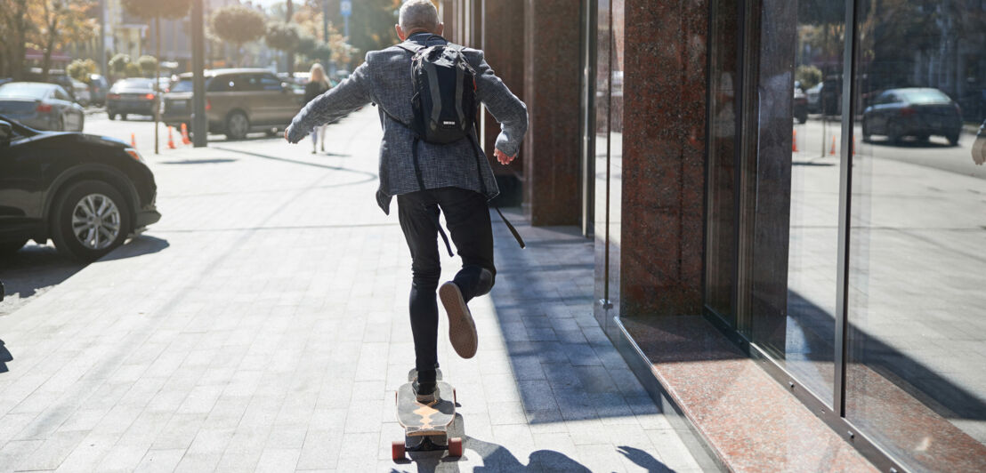 Rückansicht: Mann im Business-Look mit Rucksack fährt auf einem Longboard.