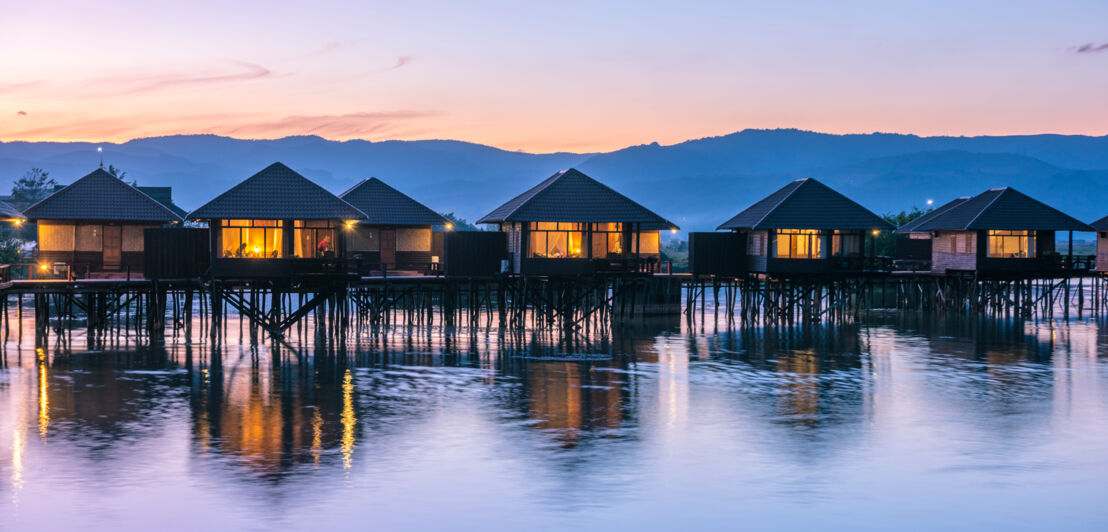 Beleuchtete Bungalows auf Stelzen im Wasser bei Sonnenuntergang