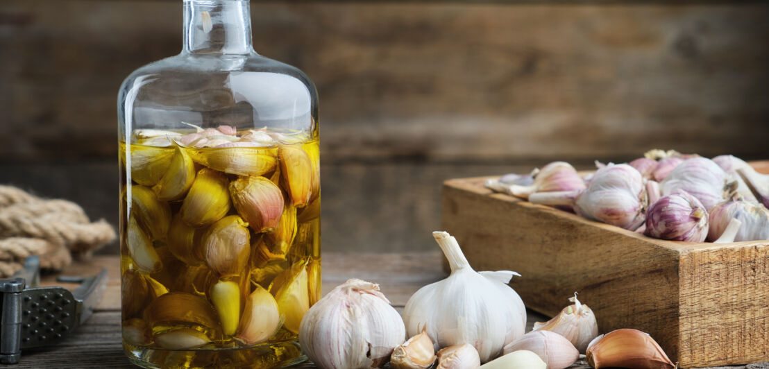 Eingelegte Knoblauchzehen in einem Glas neben einer Holzkiste mit frischem Knoblauch