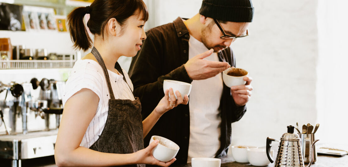 Zwei Menschen haben mehrere weiße Tassen mit Kaffee vor sich und riechen an ihnen