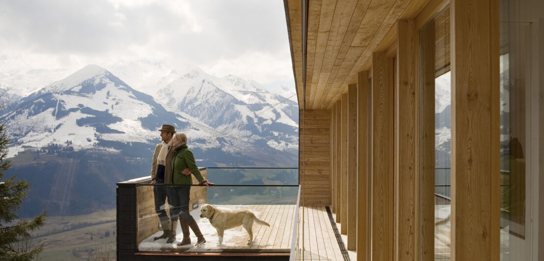 Paar mit Hund auf einer Holzterrasse eines Chalets, im Hintergrund die Berge