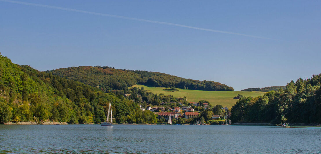 Ein See mit einem Segelschiff, dahinter hügelige Landschaft mit Wald