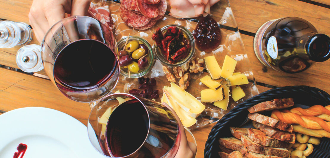 Zwei Personen stoßen mit Rotwein an, auf dem Tisch sind verschiedene Snacks verteilt