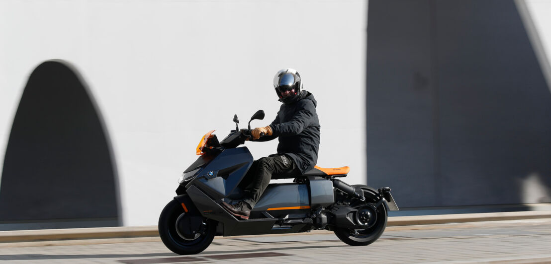 Ein grauer BMW CE 04 fährt an einer weißen Fassade vorbei, der Fahrer schaut in die Kamera.