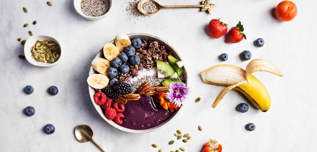 Schüssel mit frischem Obst wie Bananenscheiben, Heidelbeeren, Brombeeren, Erdbeeren, Kiwi, Nüssen und Samen.