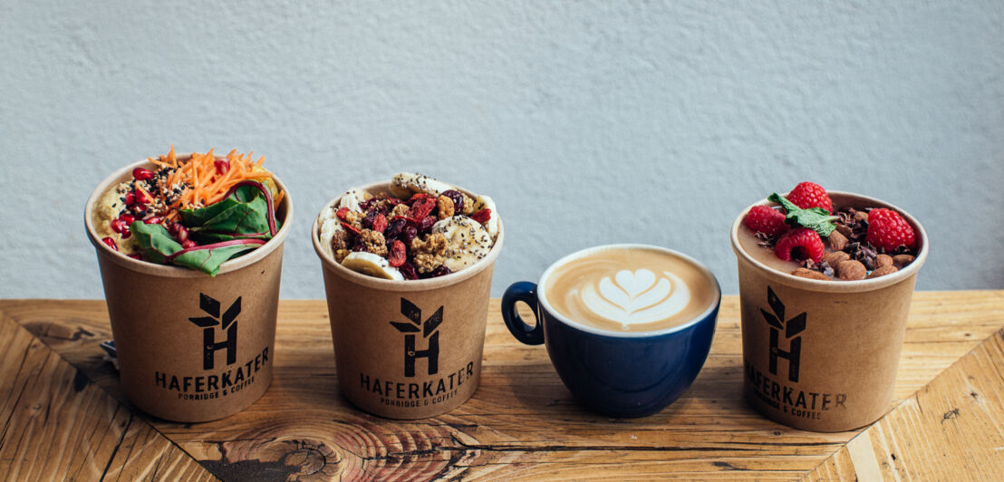 Drei mit Porridge und Obst gefüllte Becher und ein Cappuccino.