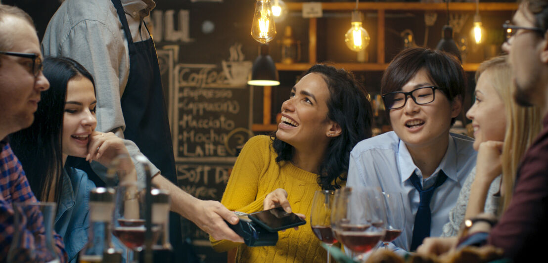 Eine junge Frau mit einer Gruppe Freunde bezahlt kontaktlos mit ihrem Smartphone an einem Tisch in einem Restaurant