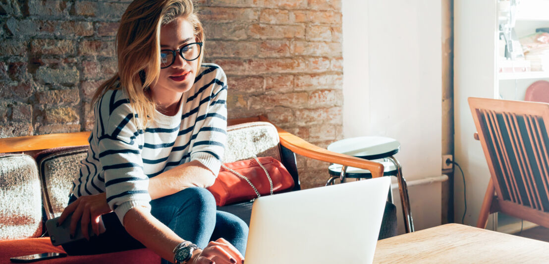 Eine junge Frau sitzt in einem Zimmer mit Vintage-Möbeln auf einem Sofa und arbeitet an ihrem Laptop