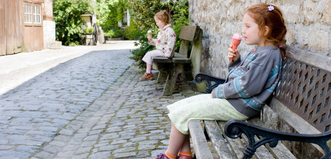 Zwei Mädchen sitzen in einer historischen Gasse jeweils auf einer Bank.