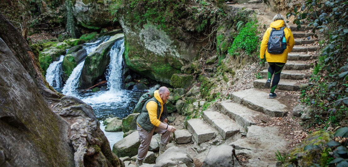 Zwei Wanderer steigen neben einem kleinen Wasserfall Steinstufen hinauf.