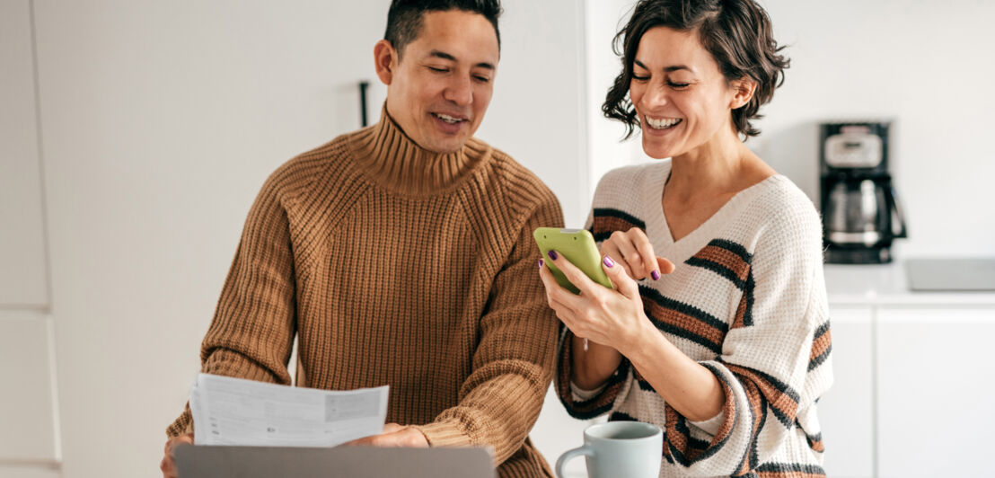 Ein Paar steht mit einer Abrechnung in der Küche und schaut gut gelaunt auf ein Smartphone