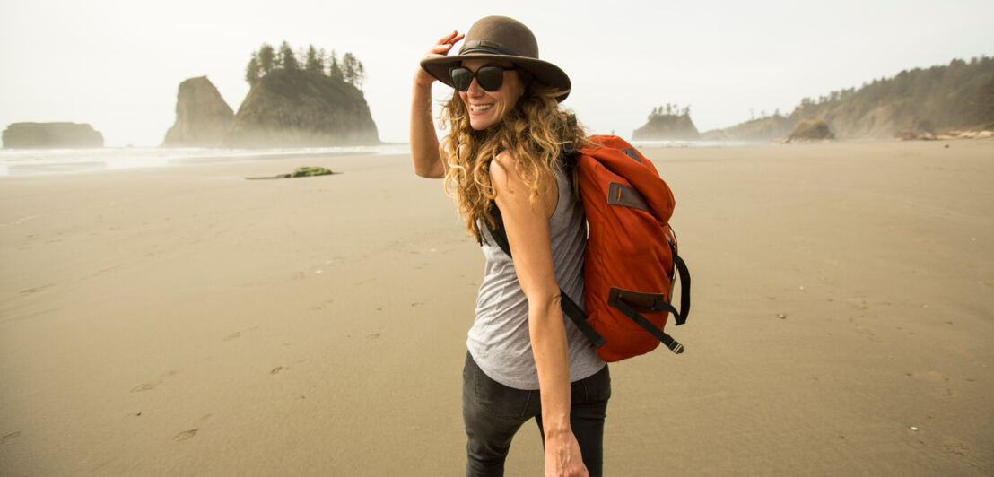 Eine gut gelaunte Frau mit Hut und Rucksack geht am Strand entlang