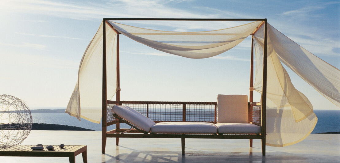 Himmelbett mit im Wind wehenden Vorhängen auf einer Terrasse am Meer