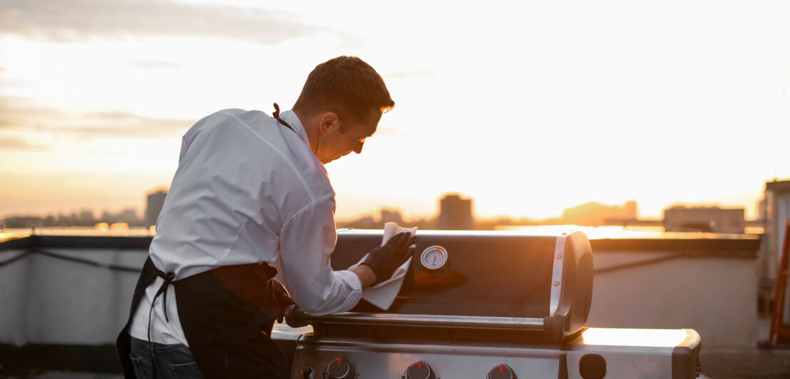 Mann in schwarzen Handschuhen und Schürze wischt im Sonnenuntergang die Grillgeräte zum Grillen ab. Sonniger Sonnenuntergang