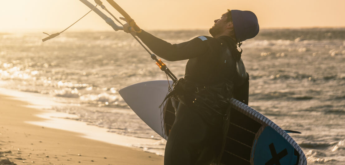 Porträt eines Wellenkitesurfers, der mit seinem Brett und einem Drachen bei wunderschönen gelben Sonnenuntergangsbedingungen am Strand aufsteigt