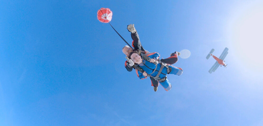 Eine Frau und ein Mann beim Tandem-Fallschirmsprung, im Hintergrund ein rotes Flugzeug