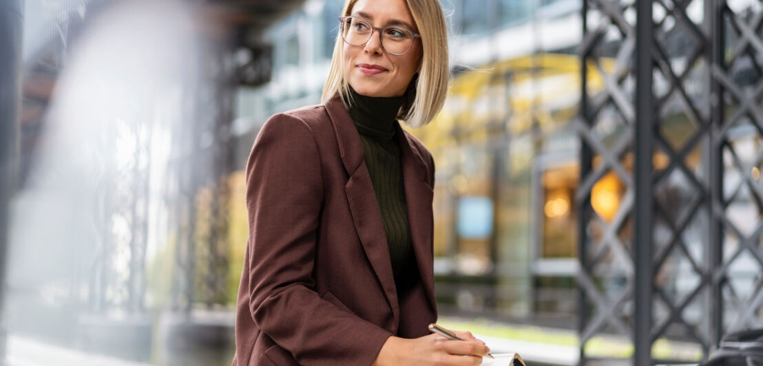 Eine Frau im Blazer sitzt mit einem Notizbuch und einem Kugelschreiber in einer industriell anmutenden Umgebung