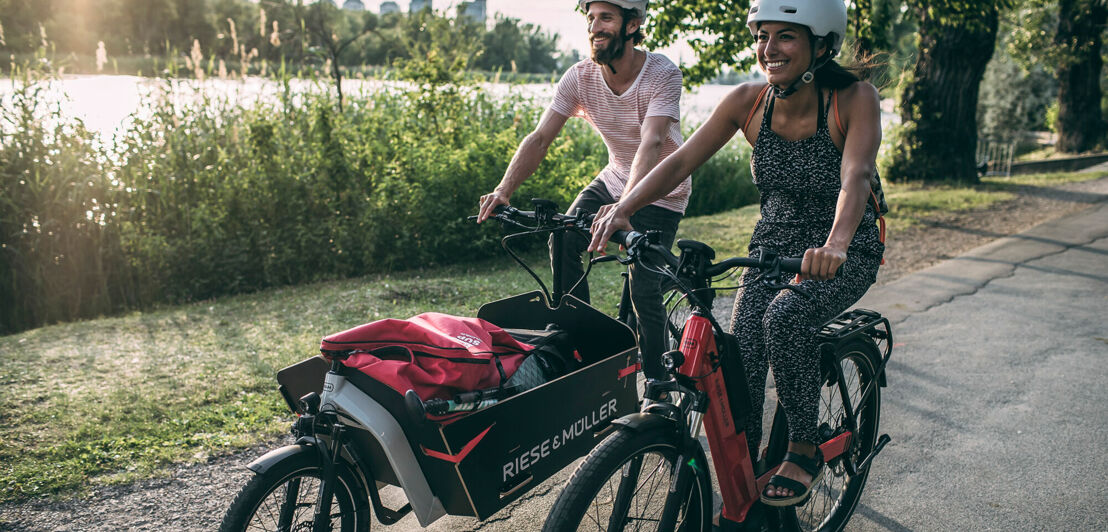 Zwei Radfahrer an einem Fluss, einer fährt ein E-Lastenrad