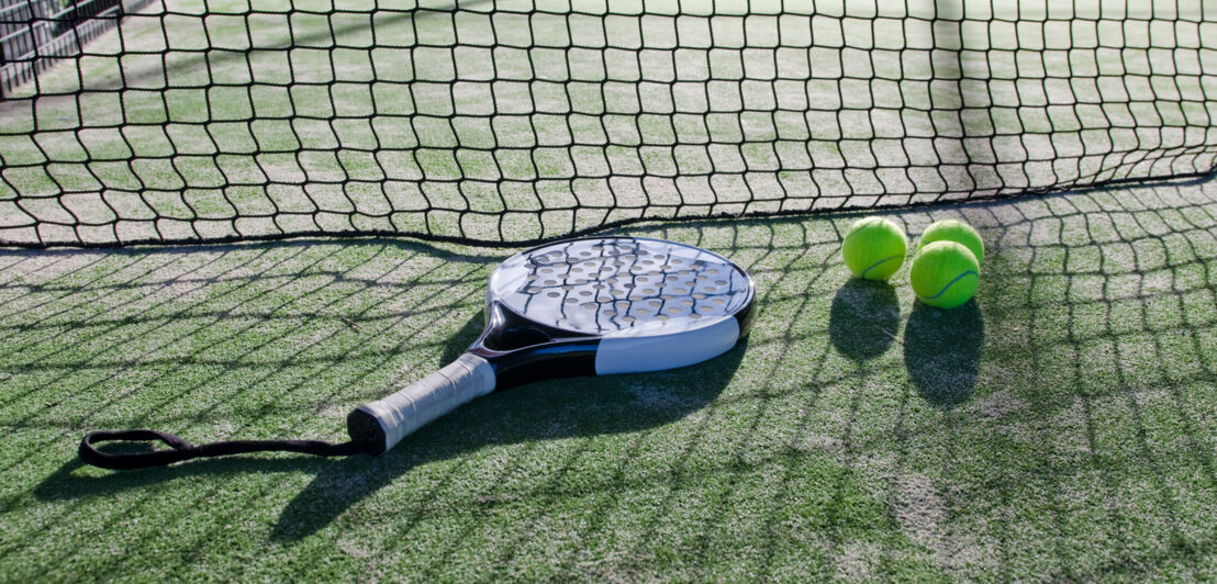 Padel-Schläger mit Löchern und Schlaufe, daneben liegen drei Tennisbälle.