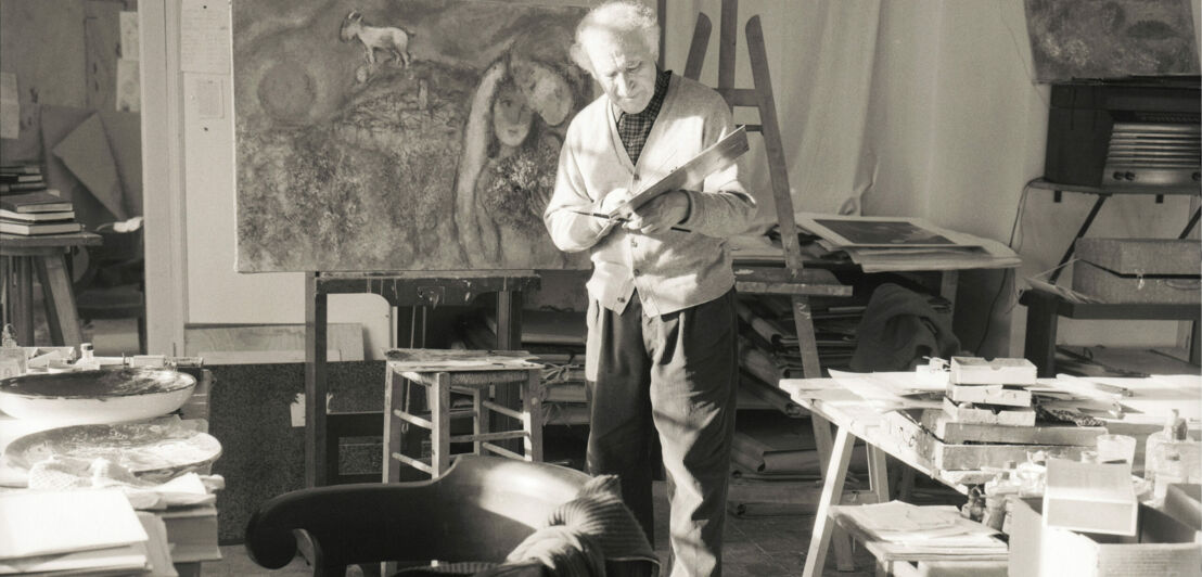 Historische Schwarzweiß-Fotografie des Malers Marc Chagall in seinem Studio vor einem Gemälde (1957)