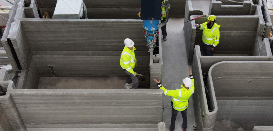 Baustelle mit Bauarbeitern inmitten den Mauern eines Hauses, das mit einem 3D-Drucker erstellt wird