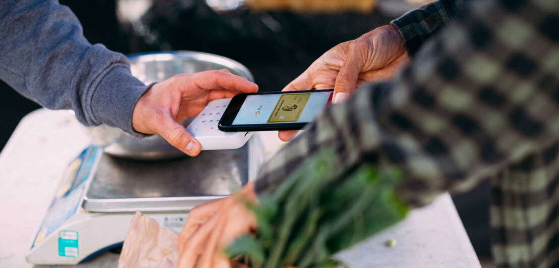 Kontaktlose Zahlung mit einer goldenen American Express Kreditkarte auf einem Smartphone, das an einem Marktstand an ein Kartenlesegerät gehalten wird