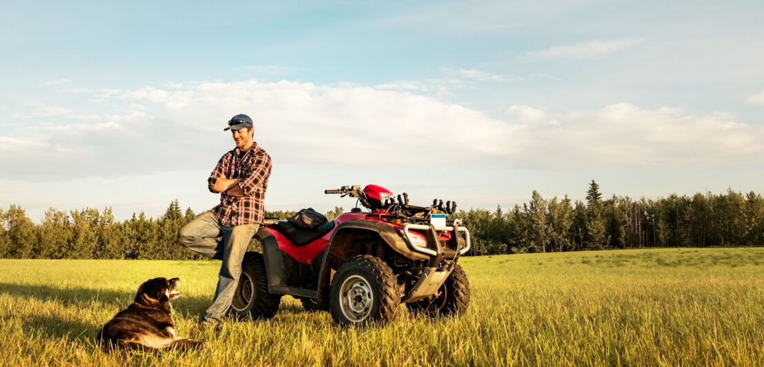 Ein Mann lehnt an einem Quad auf einem Feld, daneben ein Hund