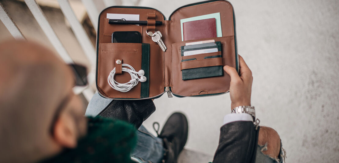 Eine sitzende Person von oben fotografiert, die eine Männerhandtasche aufklappt.