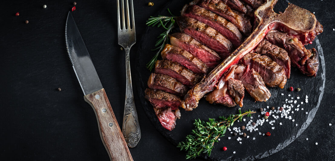 Ein gegrilltes, geschnittenes Stück Fleisch liegt auf einer Schieferplatte, daneben Messer und Gabel. 