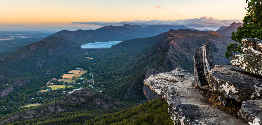 Blick in eine zerklüftete, grüne Berglandschaft mit See von einem Felsvorsprung