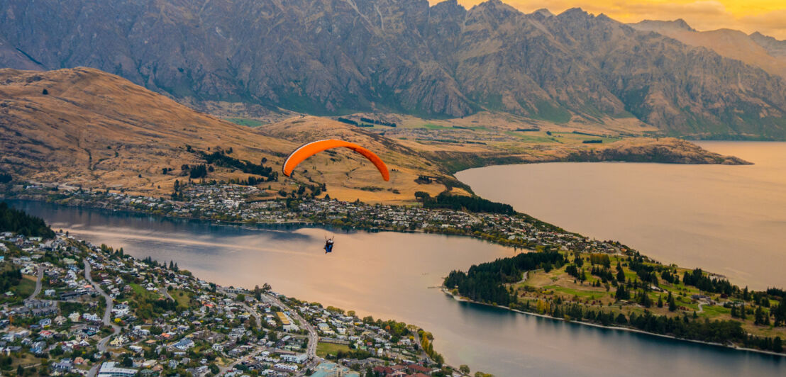 Person mit Gleitschirmflieger über einer Landschaft mit See, Stadt und Bergen