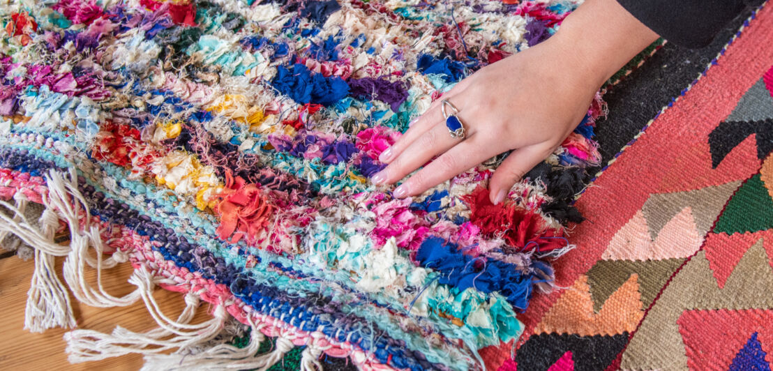 Detailaufnahme eines bunten Teppichs über den eine Hand mit blauem Ring streicht